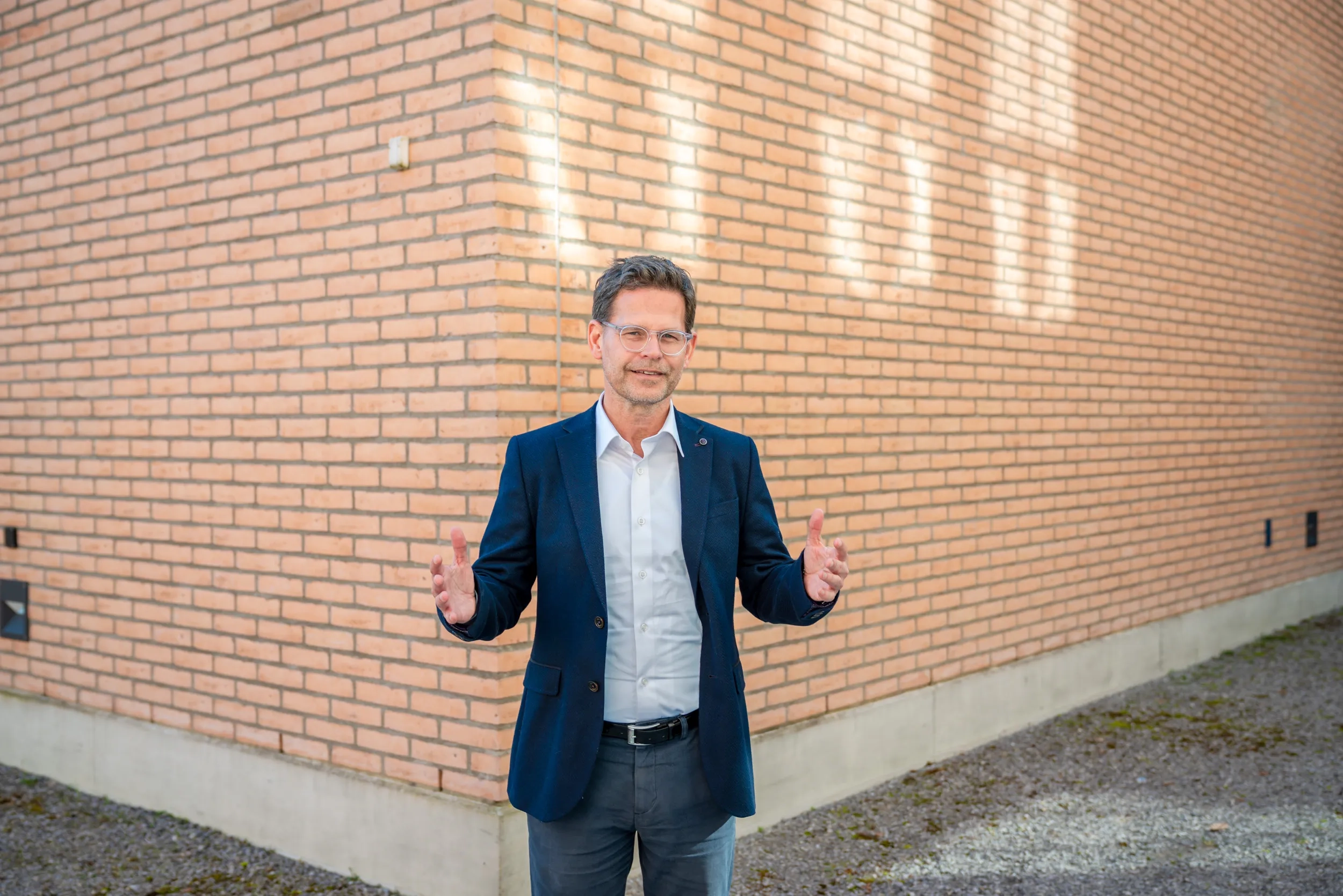 Thomas Biegel steht im Freien an der Ecke eines hellbraunen Backsteingebäudes. Er trägt eine Brille, ein weißes Hemd, ein dunkelblaues Sakko und eine graue Stoffhose mit schwarzem Gürtel. Er blickt direkt in die Kamera und hält beide Hände auf Hüfthöhe in einer offenen, einladenden Geste. Auf die Backsteinwand im Hintergrund fällt helles Sonnenlicht.