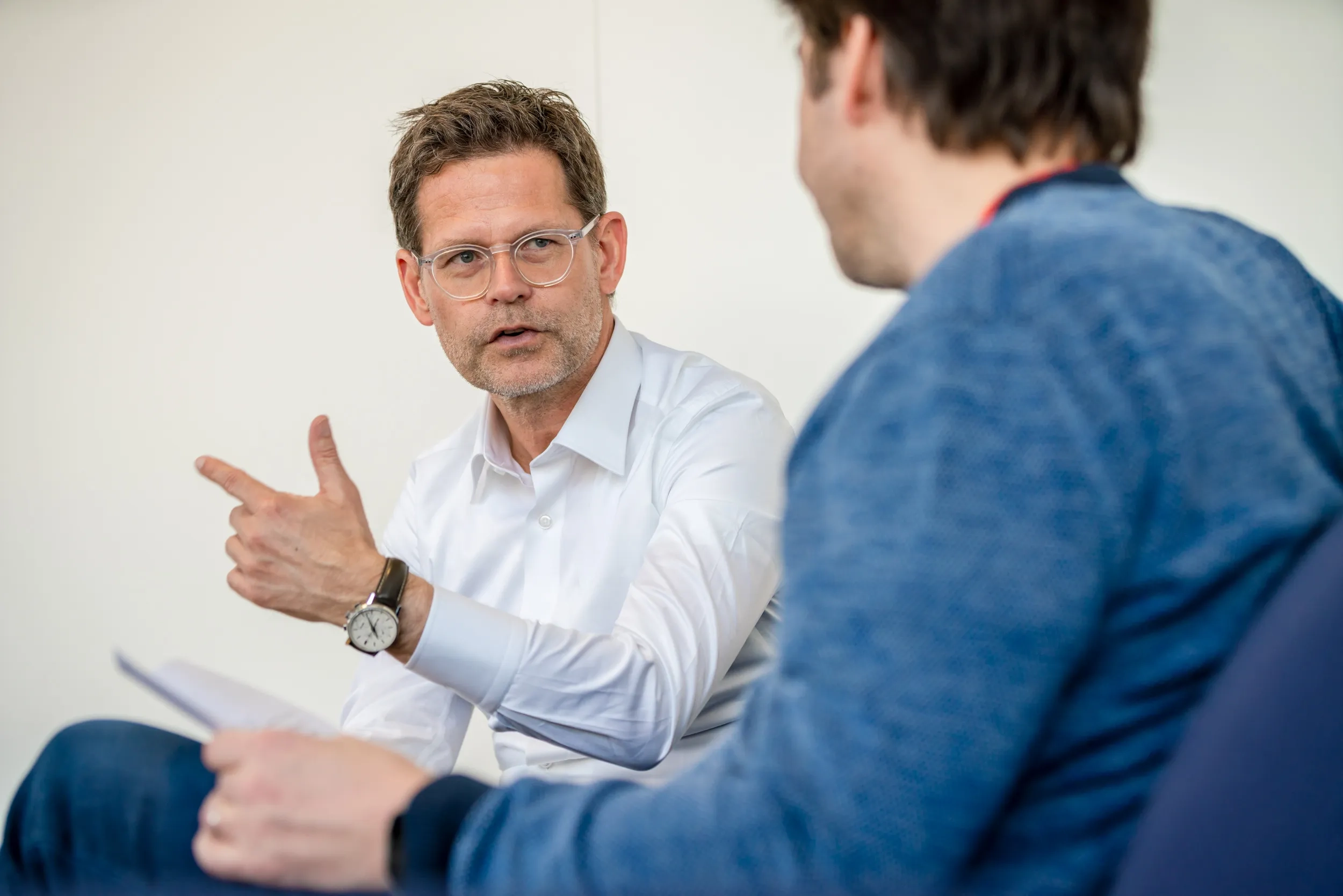 Thomas Biegel in einer aktiven Gesprächssituation im Innenraum vor einem weißen Hintergrund. Er sitzt und spricht mit einer zweiten Person, die im rechten Vordergrund unscharf und nur im Anschnitt von hinten (im blauen Pullover) zu sehen ist. Thomas Biegel trägt eine Brille und ein weißes Hemd. Er hält ein Dokument in der linken Hand und unterstreicht seine Worte mit einer zeigenden Handbewegung seiner rechten Hand. An seinem linken Handgelenk trägt er eine Armbanduhr. Er blickt seinem Gegenüber fokussiert ins Gesicht.