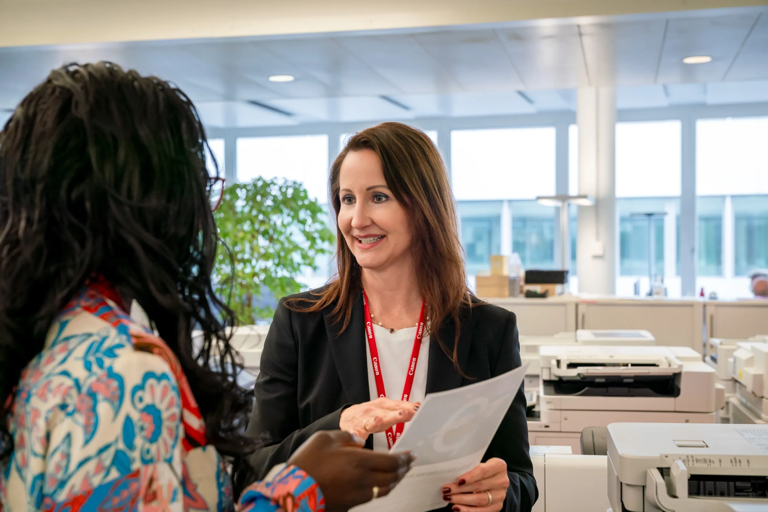 Corinne von Felten im Gespräch mit einer Mitarbeiterin im Canon-Büro: Sie hält Papiere in der Hand und lächelt; im Hintergrund ein heller Grossraum mit weiteren Canon-Geräten und einer Topfpflanze.