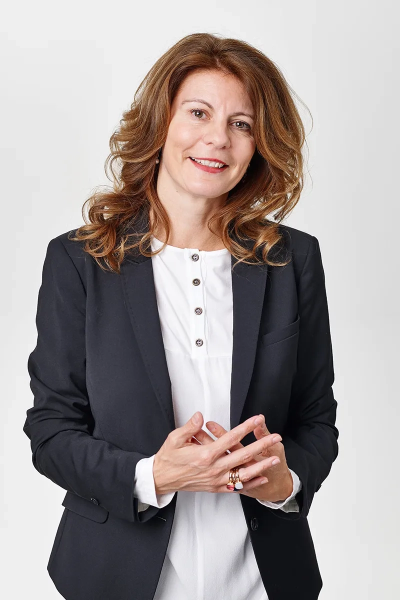 Bildbeschreibung: Ein professionelles Business-Portrait von Andrea Rutishauser von BWI vor einem neutralen, hellen Hintergrund. Erscheinungsbild: Sie hat schulterlanges, braunes, leicht welliges Haar und blickt direkt und freundlich in die Kamera. Kleidung & Pose: Sie trägt einen schwarzen Blazer über einer weißen Bluse mit Knopfleiste. Ihre Hände hält sie auf Bauchhöhe locker ineinandergelegt. An der rechten Hand trägt sie einen auffälligen Ring. Der Gesamteindruck des Bildes ist professionell und nahbar.