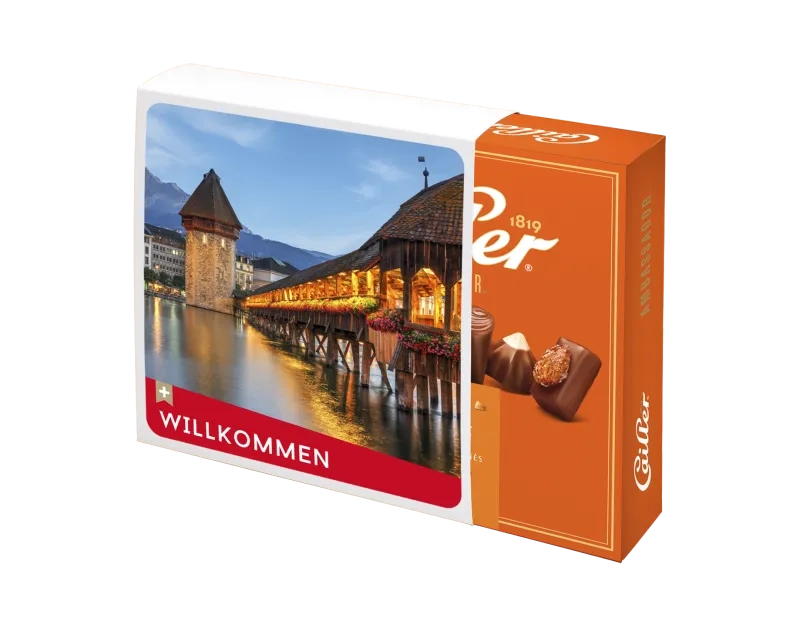 Eine orangefarbene Cailler Ambassador Pralinenschachtel vor schwarzem Hintergrund. Über die linke Hälfte der Schachtel ist ein bedruckter Schuber geschoben. Dieser zeigt ein Foto der beleuchteten Luzerner Kapellbrücke bei Abenddämmerung. Im unteren Bereich des Schubers befindet sich ein roter Balken mit einem weißen Schweizerkreuz und dem Wort "WILLKOMMEN".