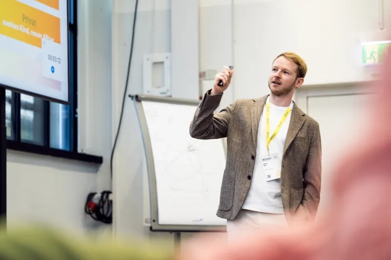 Porträt eines Mannes mit braun-meliertem Sakko und gelbem Lanyard. Er hält ein Gerät in der Hand, zeigt nach oben und spricht, während links ein Präsentationsbildschirm und im Hintergrund ein Flipchart zu sehen sind.