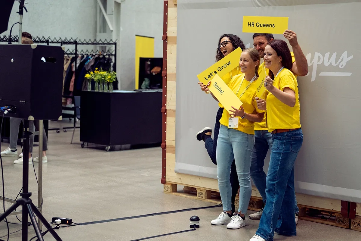 Vier Personen in gelben T-Shirts posieren an einer Fotobox; sie halten gelbe Schilder mit «Let’s Grow», «Smile!» und «HR Queens»; lockere Event-Atmosphäre im Hintergrund.