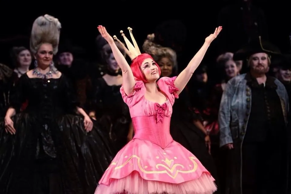 Die Opernsängerin Erin Morley als Olympia auf der Bühne der Metropolitan Opera in New York (2015). Sie steht im Rampenlicht und trägt ein leuchtend pinkfarbenes Kleid mit Puffärmeln, einem Tüllrock und goldenen Verzierungen. Auf dem Kopf trägt sie eine auffällige rote Kurzhaarperücke und eine große, goldene Krone. Mit stark betontem, puppenhaftem Make-up und einem strahlenden Lächeln reißt sie die Arme triumphierend in die Höhe. Im abgedunkelten Hintergrund stehen weitere Darsteller in aufwendigen, überwiegend dunklen historischen Kostümen.