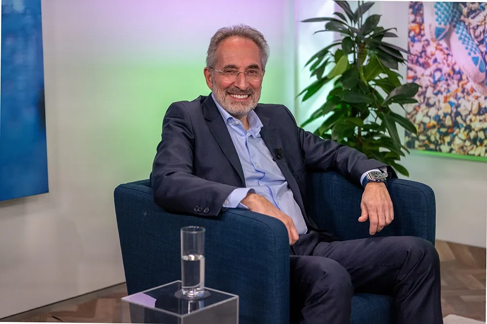 Frédéric Patitucci sitzt entspannt in einem blauen Sessel und lächelt in die Kamera. Er trägt einen dunkelblauen Anzug mit hellblauem Hemd und hat ein Mikrofon am Revers. Im Hintergrund sind eine grüne Zimmerpflanze, farbige Wandbeleuchtung und ein modernes Kunstwerk zu sehen. Auf einem gläsernen Beistelltisch vor ihm steht ein Glas Wasser.