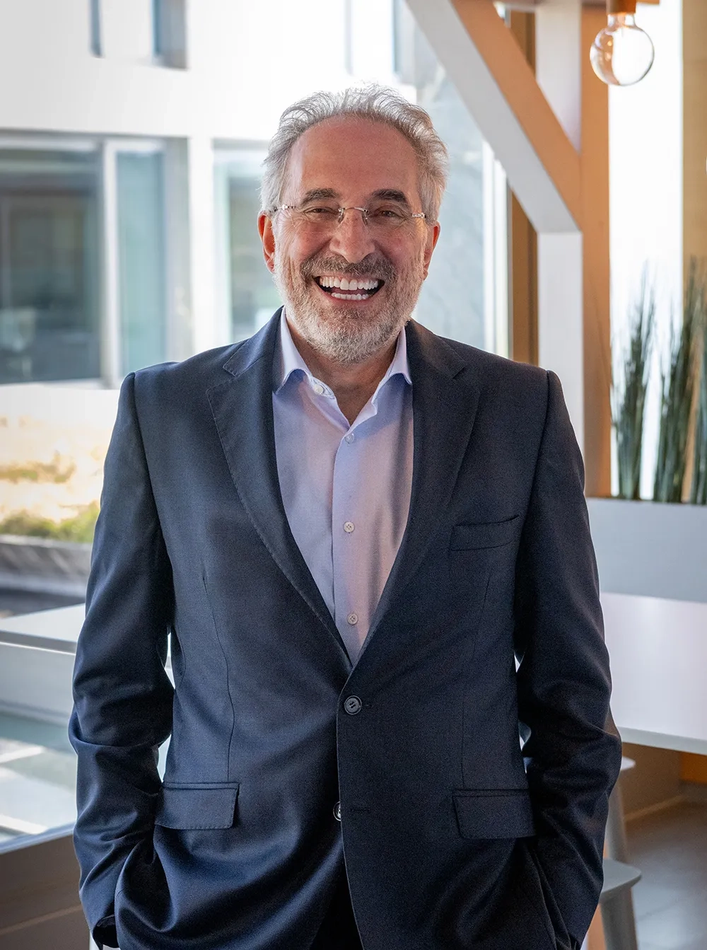 Frédéric Patitucci steht lächelnd in einem modernen, lichtdurchfluteten Büroraum. Er trägt einen dunkelblauen Anzug mit offenem Hemd ohne Krawatte und hat die Hände locker in den Hosentaschen. Im Hintergrund sind grosse Fenster und Pflanzen zu sehen.