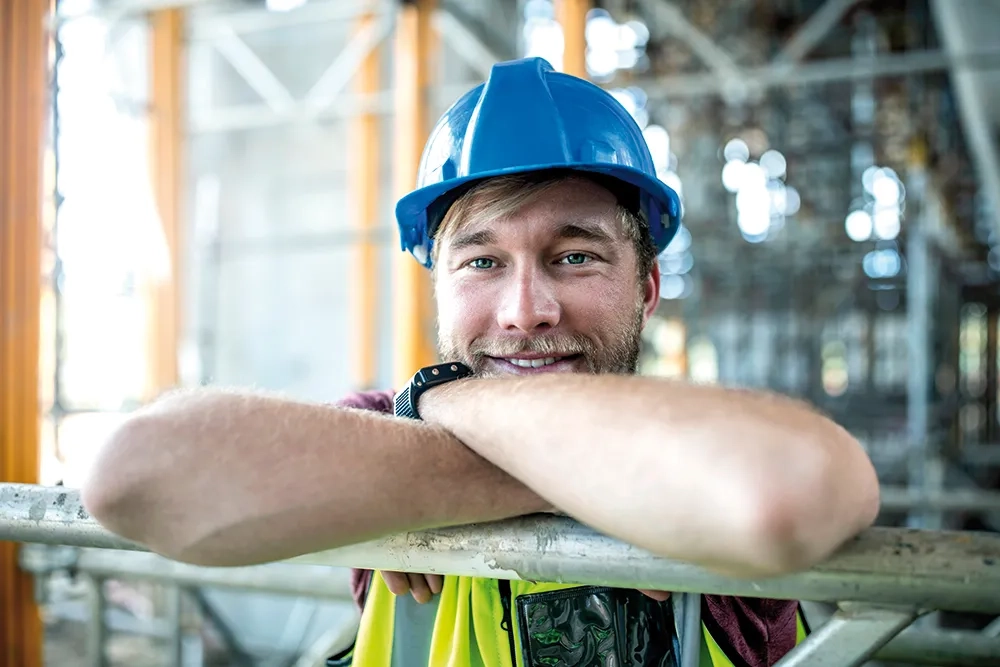 Auf einer Baustelle lehnt eine Person im blauen Helm entspannt am Geländer und lächelt; Gerüst im Hintergrund.