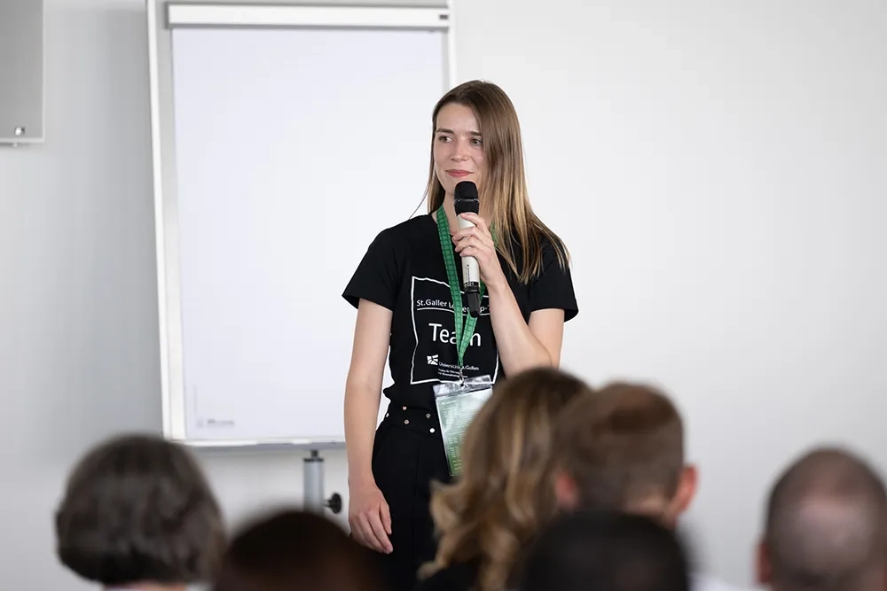 Eine Frau mit langen braunen Haaren steht vor einem weißen Flipchart und spricht in ein Handmikrofon. Sie trägt ein schwarzes T-Shirt mit dem Aufdruck „Team“ und ein grünes Schlüsselband mit einem Ausweis. Im unscharfen Vordergrund sind die Hinterköpfe von sitzenden Zuschauern zu sehen.
