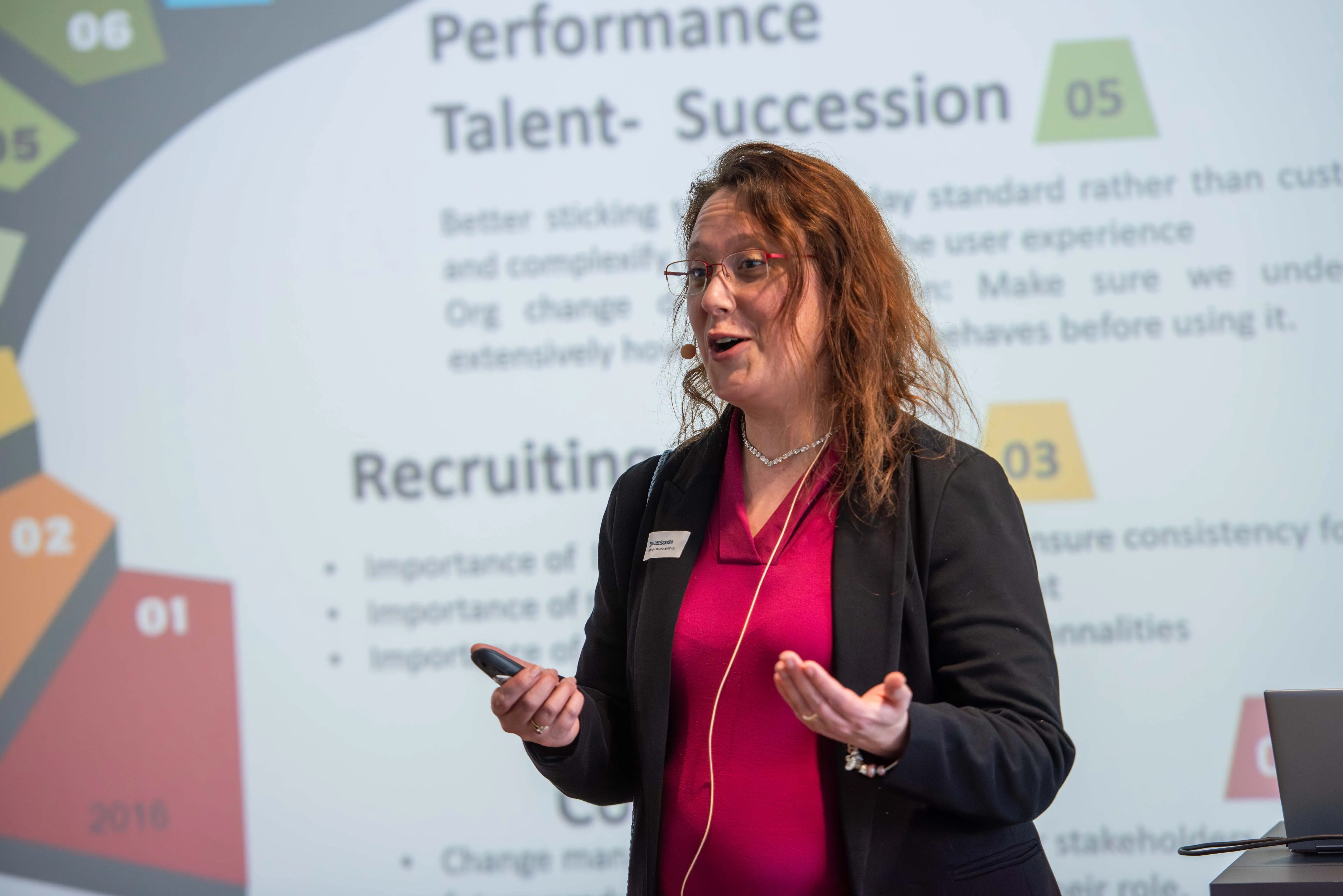 Lynn Van Oossanen, eine Frau mit Brille und Headset-Mikrofon hält eine engagierte Präsentation. Sie trägt einen pinkfarbenen Blazer über einem dunklen Oberteil. Im Hintergrund ist eine Präsentationsfolie mit den Titeln „Performance“, „Talent-Succession“ und „Recruiting“ zu sehen.