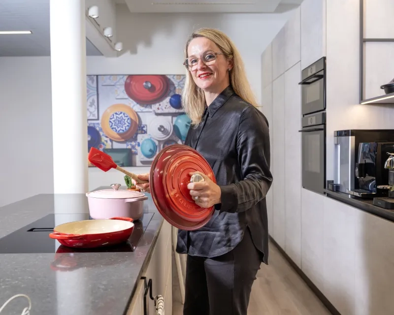 Maryline Tissot im schwarzen Hemd steht an einem Kochfeld in einer hellen Küche. Sie blickt lächelnd in die Kamera und hält einen roten Le-Creuset-Deckel sowie einen roten Silikon-Spatel in den Händen. auf dem Kochfeld stehen weitere Bunte Töpfe und Pfannen der Marke.