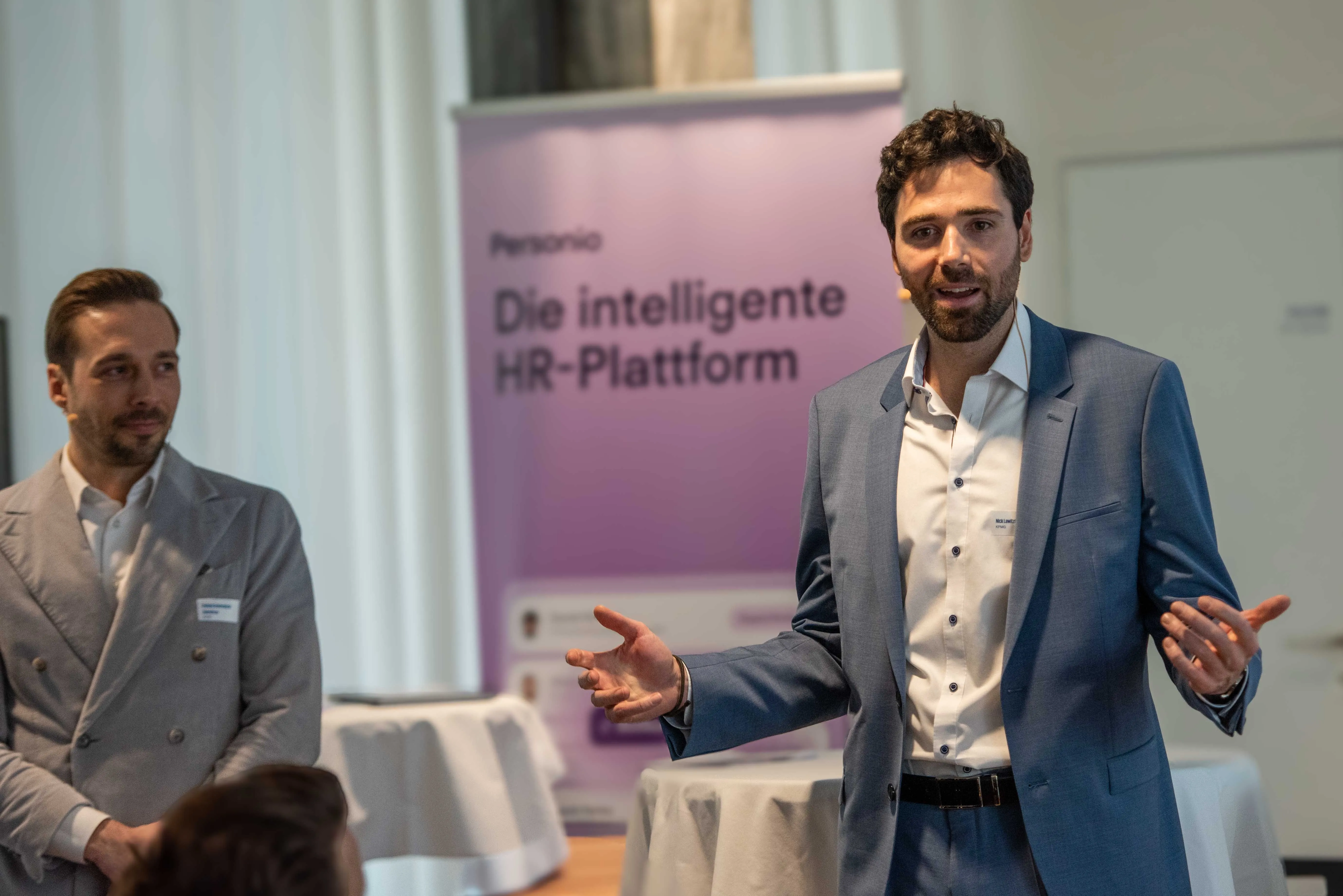 Zwei Männer in Business-Kleidung bei einem Vortrag. Der Mann im Vordergrund, Nick Lawitzke, trägt ein blaues Sakko und spricht gestikulierend in den Raum. Dahinter steht Patrick Schwesig im grauen Sakko. Im Hintergrund steht ein lila Roll-up-Banner mit der Aufschrift „Personio – Die intelligente HR-Plattform“.