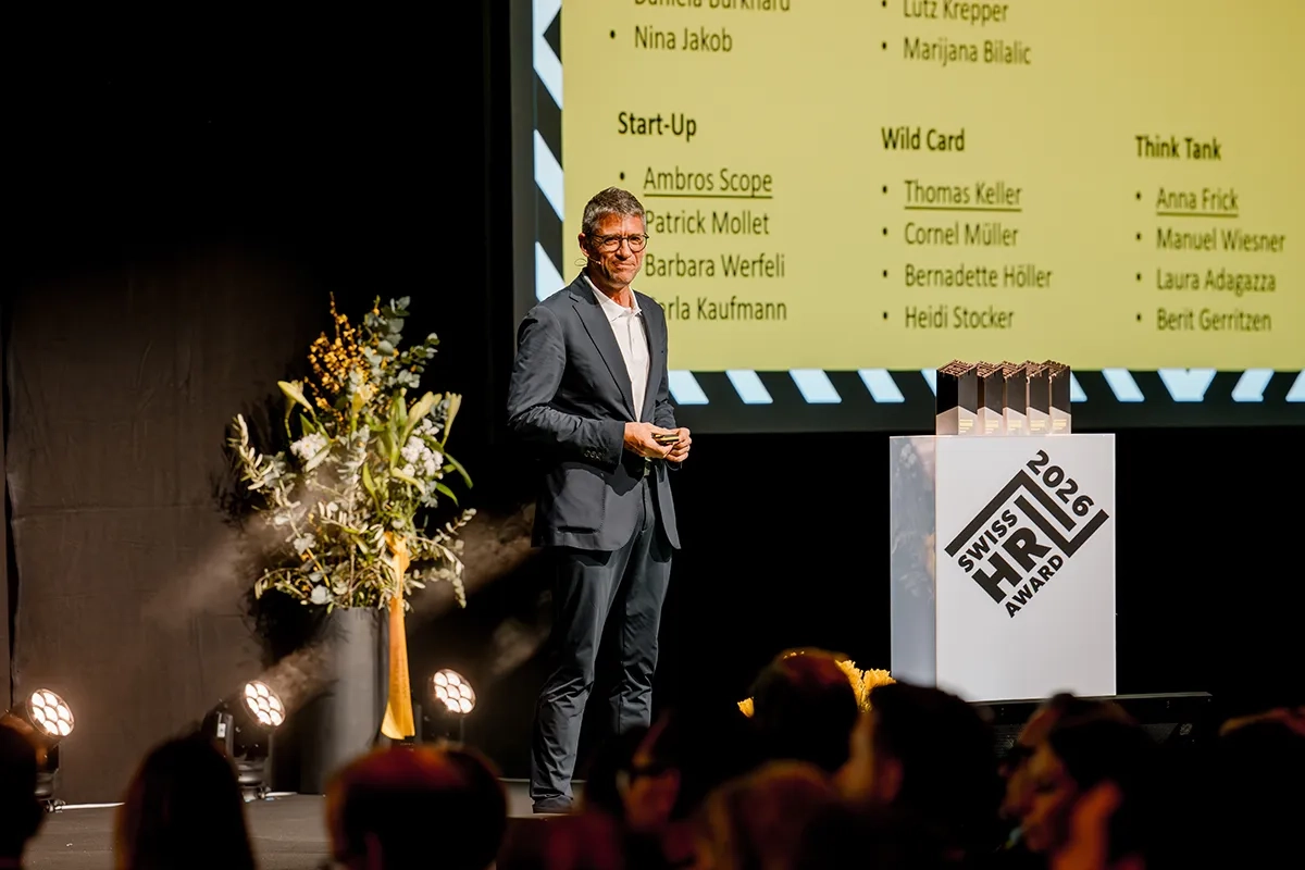 Pascal Scheiwiller, Jury-Präsident, steht im dunklen Anzug auf der Bühne des Swiss HR Award 2026 und hält einen Vortrag. Rechts neben ihm steht ein weißes Podest mit dem Award-Logo und mehreren schwarzen Trophäen. Im Hintergrund wird eine gelbe Präsentationsfolie mit verschiedenen Kategorien und Namen eingeblendet, während im unscharfen Vordergrund das Publikum zu sehen ist.