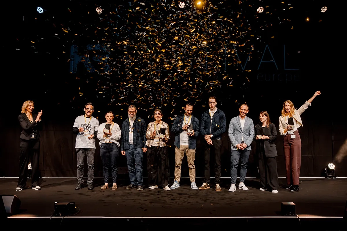Eine Gruppe von glücklichen Gewinnern steht mit ihren schwarzen Trophäen auf einer Bühne, während goldenes Konfetti von der Decke regnet. Links im Bild steht eine Moderatorin, die den Gewinnern applaudiert.