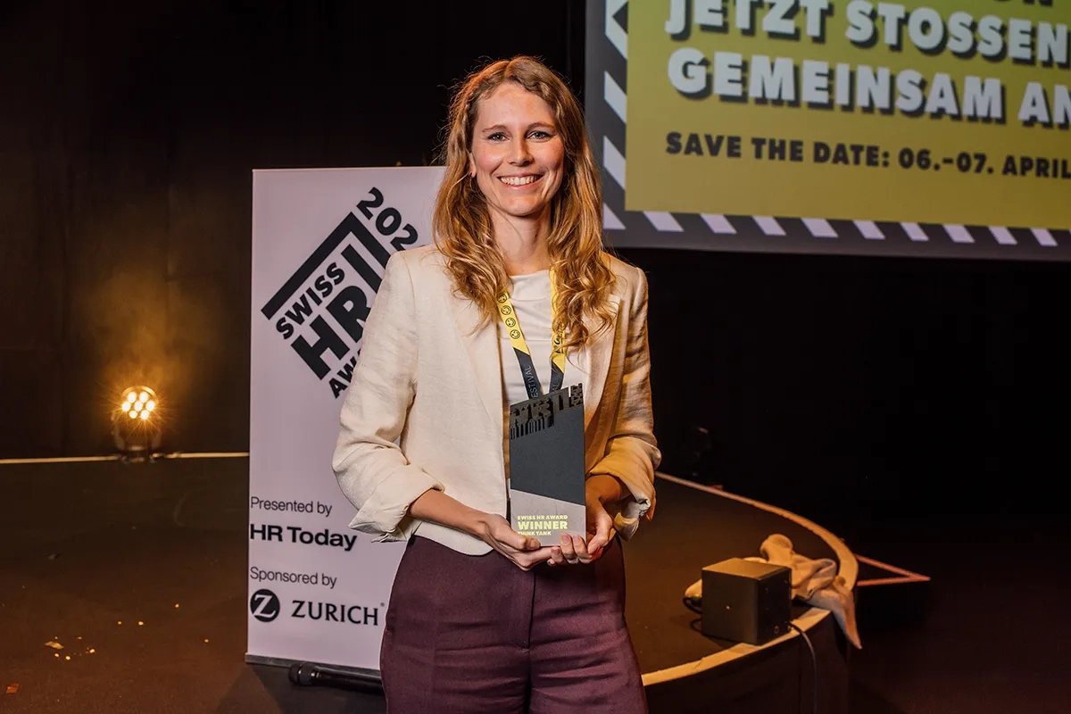 Eine lächelnde Frau mit langen blonden Haaren steht auf einer Bühne und präsentiert ihre Trophäe in die Kamera. Auf der Trophäe steht "Swiss HR Award Winner Think Tank". Neben ihr steht ein weißer Aufsteller mit der Aufschrift "Swiss HR Award 2026"
