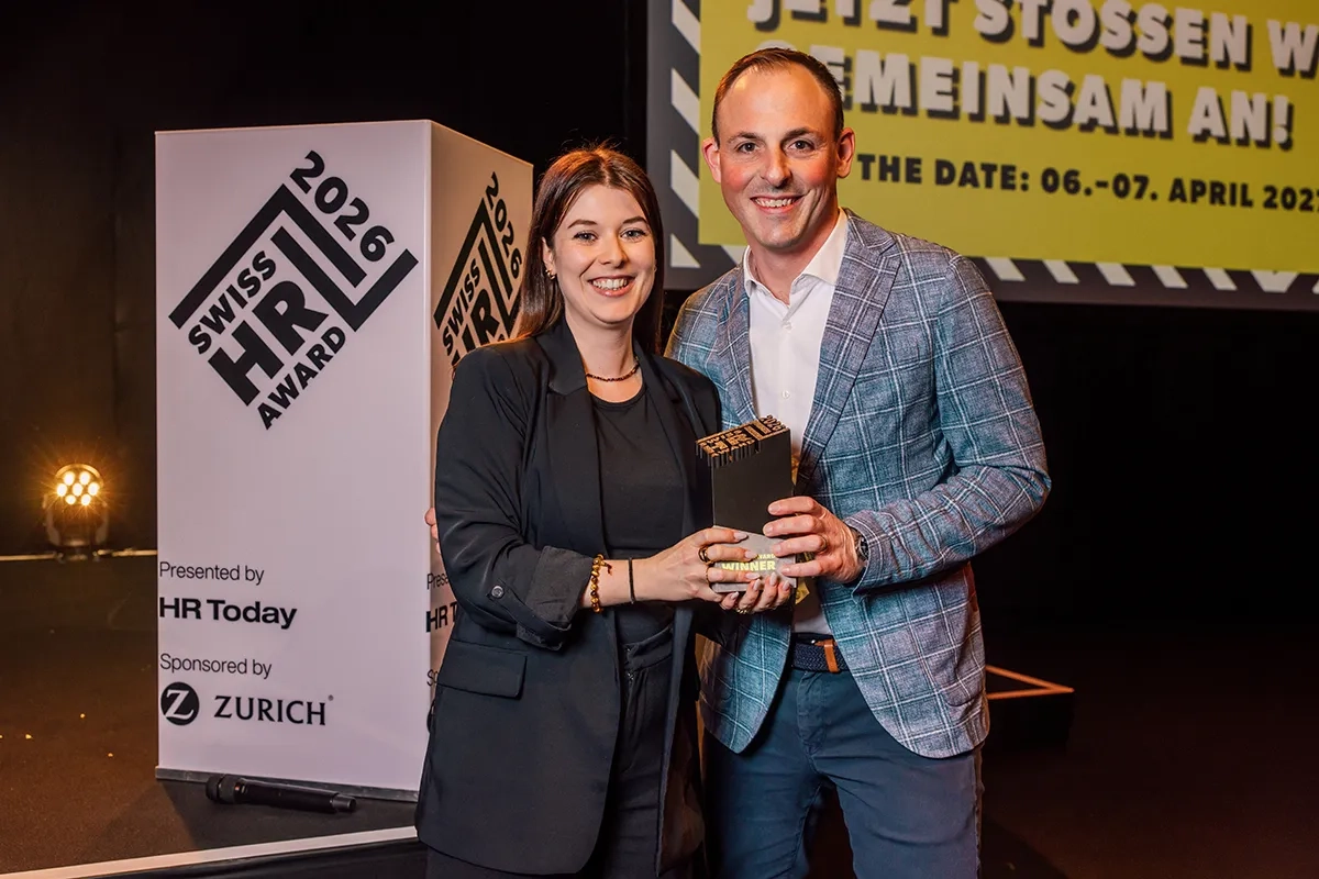 Eine Frau im schwarzen Blazer und ein Mann im blau karierten Sakko stehen gemeinsam auf der Bühne und halten strahlend eine schwarze Trophäe des Swiss HR Award in den Händen.