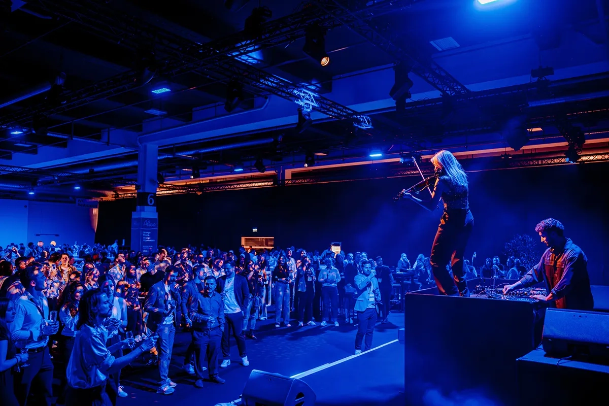 Die Musikerin Laura Zimmermann spielt E-Geige auf einer kleinen Bühne während der Networking-Party des HR FESTIVAL europe 2026. Der Raum ist in stimmungsvolles, tiefblaues Licht getaucht. Neben ihr steht ein DJ an seinem Pult, während im Vordergrund das Publikum mit Getränken in der Hand feiert, sich unterhält und der Musik lauscht.