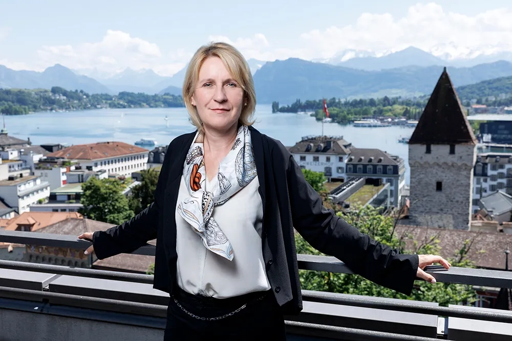 Nathalie Leschot von der Suva steht auf einer Terrasse mit Blick auf die Stadt Luzern. Im Hintergrund sind der Vierwaldstättersee, historische Gebäude und die Alpen zu sehen. Sie schaut selbstbewusst in die Kamera und hält sich mit beiden Händen am Geländer fest.
