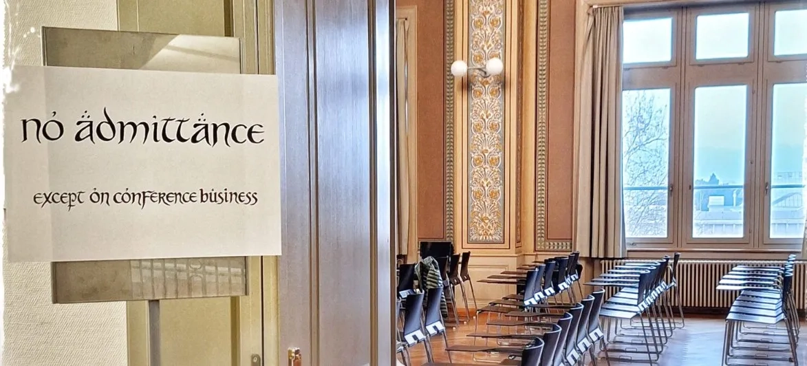 Das Bild zeigt den Blick durch eine geöffnete Tür. Im Vordergrund auf der linken Seite ist ein weisses Papierschild an einer Scheibe angebracht. Darauf steht in einer an das "Herr der Ringe"-Universum erinnernden Schriftart: "no admittance" und darunter etwas kleiner "except on conference business ". Viele Buchstaben sind dekorativ mit Akzenten oder Umlauten versehen. Der Spruch ist eine humorvolle Abwandlung des Schildes an Bilbo Beutlins Tür ("No admittance except on party business"). Auf der rechten Bildhälfte blickt man durch den hölzernen Türrahmen in einen grossen Konferenzraum in einem älteren, herrschaftlichen Gebäude. Die Wände sind mit klassischen, floralen Ornamenten verziert und eine runde Wandlampe ist zu sehen. Im Raum stehen ordentlich aufgereihte, leere moderne Konferenzstühle in Schwarz, die mit kleinen seitlichen Schreibflächen ausgestattet sind. Hohe Fenster lassen helles Tageslicht in den Raum fallen.